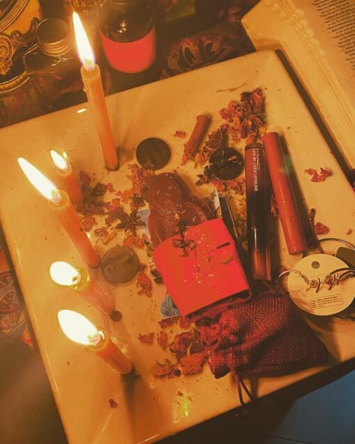 A ritual setup with lit candles, scattered herbs, lipstick, and a red pouch on a tray, surrounded by spiritual items for an attraction spell